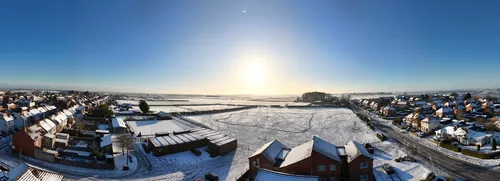This landscape panorama captures an eastward view of Bolsover in winter, just before 10 in the morning. Snow blankets both the urban residential areas and the expansive fields visible in the background, highlighting the seasonal contrast between the built environment and the open countryside. The photograph shows the rooftops of houses and other urban structures, while the sun rises low on the horizon, casting a bright light across the winter fields. The image includes the distinct separation between Bolsover's townscape and the snow-covered rural fields, resulting in a striking visual display characteristic of winter mornings.